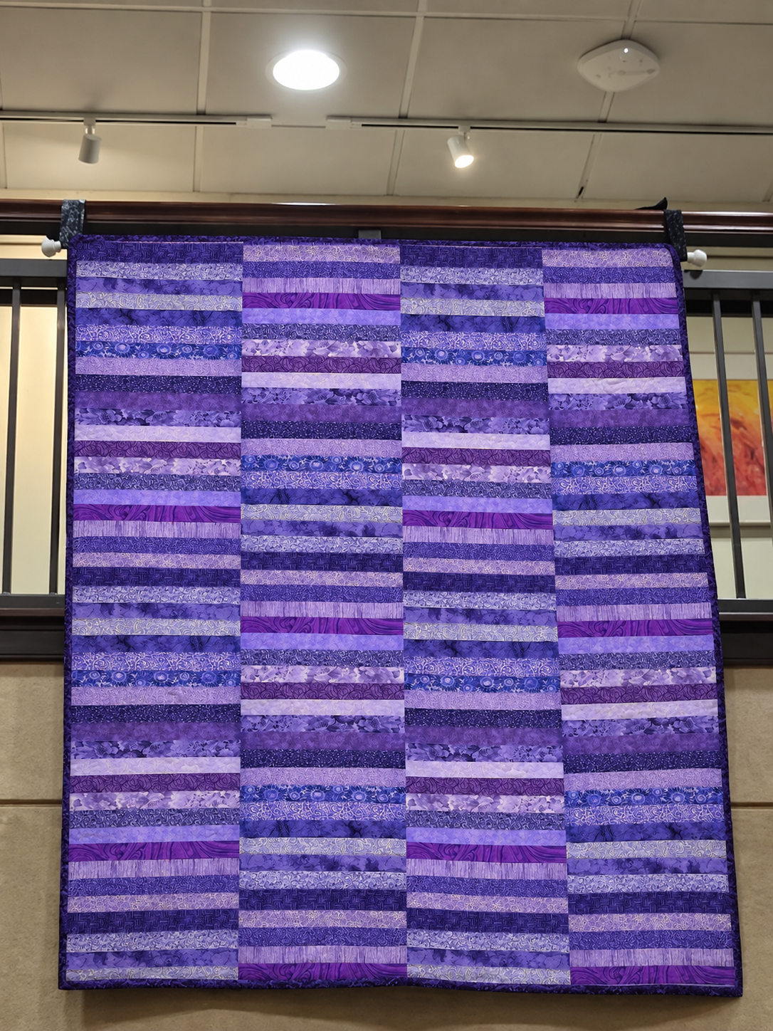 A rectangular quilt or fabric panel with horizontal stripes in various shades of purple and blue, hanging from a dark rod in an indoor space with a ceiling and window visible.
