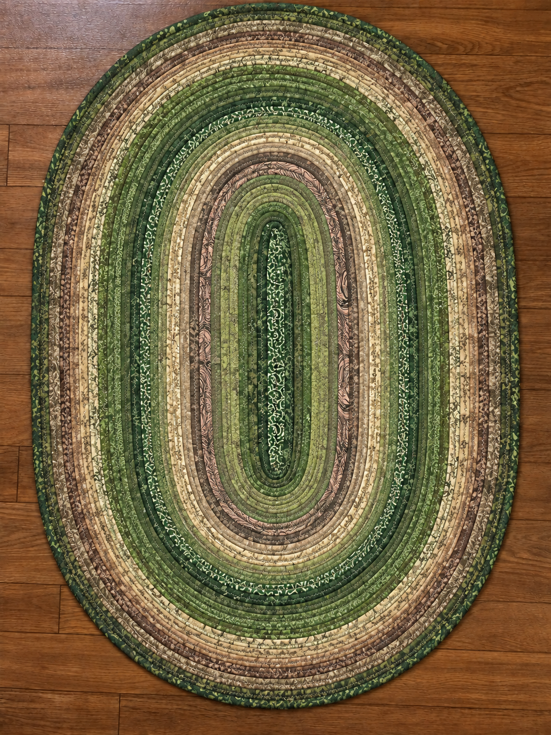 An oval braided rug with concentric bands of green, olive, beige, and brown, shown from a top-down view on a wooden floor.