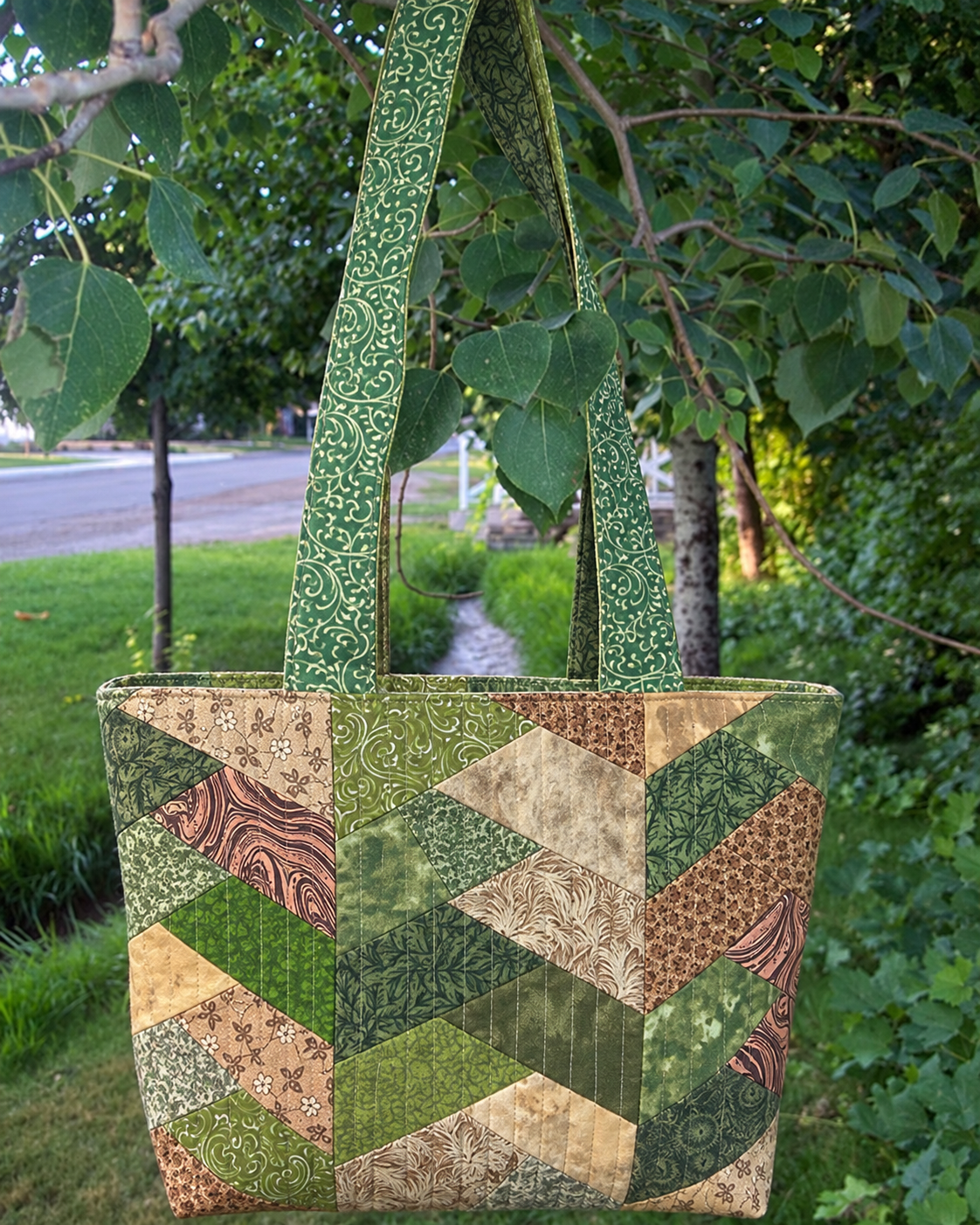 A handmade patchwork fabric tote bag hanging from a tree branch outdoors. The bag features a geometric chevron pattern made of various green, brown, and beige patterned fabric squares, with two long green patterned shoulder straps.